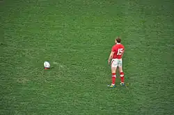 Photo d'une homme debout, de dos, immobile, s'apprêtant à frapper un ballon de rugby place quelques mètres devant lui.