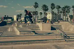 Photo d'un skater en train d'effectuer une figure de skate à Venice Beach. Il est pris en l'air durant un saut à 1 à 2&nbsp;mètres de hauteur et son skate effectue des rotations en dessous de lui. Le décor est principalement composé d'un multitude d'escaliers en béton de quelques marches. En arrière-plan assez loin, il y a des skaters et beaucoup plus loin, on aperçoit des cocotiers, la plages et des immeubles.