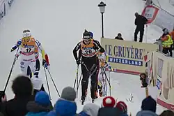 Deux skieuses de fond de face dans une montée, avec au premier plan des têtes de spectateurs, de dos.