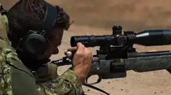 Photographie couleur. Visage de profil orienté vers la droite d'un militaire équipé d'un casque anti-bruit et épaulant, allongé, avec un fusil à lunette. De sa main droite, l'homme tire la culasse de son arme et en éjecte une douille