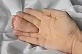 Plante de pied d'un nourrisson âgé d'un mois, maintenu entre le majeur et l'annulaire d'une main adulte, pour comparaison de taille, sur un drap blanc, à Don Det, Si Phan Don, Laos. Septembre 2020.