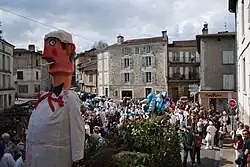 En premier plan, le mannequin Bufador et le bûcher sont remorqués par un tracteur sur une place de Nontron. Les Soufflaculs et les spectateurs sont situés tout autour.
