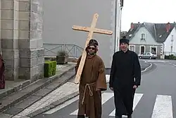 Deux hommes sont déguisés. L'un est habillé d'une robe traditionnelle de moine et de lunettes de soleil, portant sur son dos une grande croix en bois. Le deuxième porte une soutane noire, des moustaches de chat sont maquillées sur son visage.