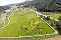Photo couleur d'une vallée comprenant plusieurs rizières. Dans l'une d'elles, au premier plan, des plants de riz de différentes couleurs (vert, jaune et noir) dessinent un cheval ailé, au-dessus d'un message en coréen.
