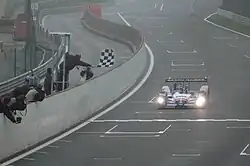 Photographie d'une voiture bleue, blanche et rouge, de face, sur une piste, devant un drapeau à damier