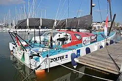 Voilier blanc amarré au ponton, vu de trois quarts arrière. Pont bleu, rouf rouge.