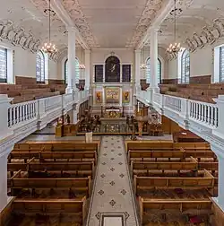 L'intérieur d'une grande salle aux murs blancs, un balcon de chaque côté. Des rangées de bancs en bois marron. Quelques reflets or au niveau du choeur.