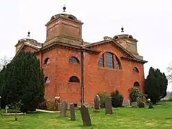 Photo d'un bâtiment en briques rouges aux murs imposants, situé au milieu d'une pelouse jonchée de pierre tombales