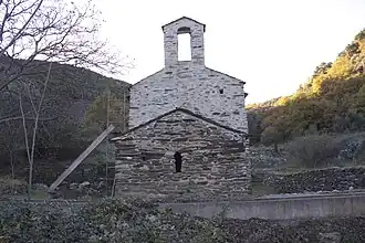 Mur en pierres percé d'une petite fenêtre en arc, surmonté d'un toit. Plus haut derrière, autre mur surmonté d'un clocher-mur.