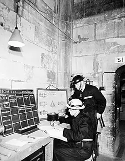 Vers la même époque, dans une salle de contrôle (où l'on distingue, contre le mur, un plan détaillé de la cathédrale) sont coordonnés des guetteurs prêts à parer aux éventuels départs d'incendies pouvant toucher le monument pendant les attaques aériennes.