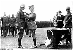 Photographie en noir et blanc d'un homme en uniforme avec une casquette à visière épinglant une médaille sur la veste d'un second homme en uniforme portant un casque de protection.