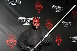 Homme maquillé en rouge et noir avec des vêtements noirs et un sabre.