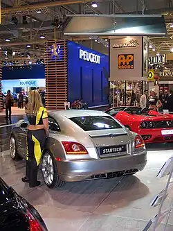 Une voiture 2 portes grise vue de l'arrière gauche exposée dans un salon aux côtés d'autres automobiles. Une femme s'appuie sur la portière conducteur de la voiture.