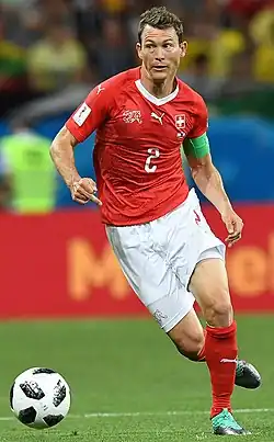 Photographie couleur d’un footballeur, de pied, en mouvement, le visage très expressif, vêtu d’un maillot rouge