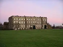 Au milieu d'une pelouse, façade d'un vaste bâtiment de trois étages sur rez-de-chaussée surélevé. 15 fenêtres par étage, un toit en terrasse bordée de colonnettes.