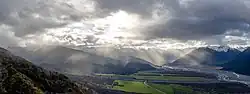 Une grande vallée où des rayons du Soleil sortis des nuages illuminent des champs et le cours d'eau ; tout est couronné par une chaîne de montagnes.