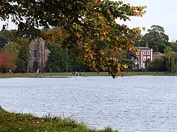 Panorama d'un lac calme en automne, avec des arbres aux couleurs jaunes et oranges. Tour médiévale et maison de briques rouges en arrière-plan. Ciel gris, atmosphère paisible, typique d'un paysage de campagne automnale.
