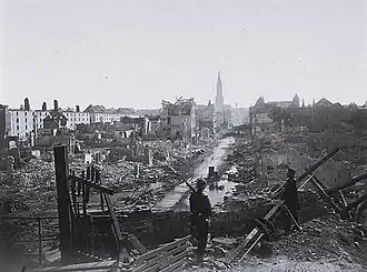 Photographie montrant une vue d'ensemble de la ville en ruine, observée par deux soldats au premier plan.