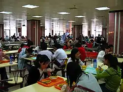 Dans une très grande salle d’architecture moderne, parsemée de massifs piliers carrés bordeaux et blancs, des étudiants sont assis à des tables pour 4 personnes et y mangent ou y discutent. Dans un angle de la salle, une TV est allumée.