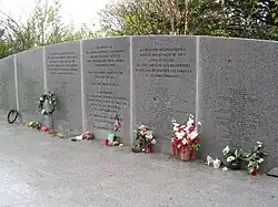 Photo centrée sur le mur en pierre commémoratif. De nombreux bouquets de fleurs et couronnes sont présents à son pied, sur le sol en pierre et il comporte des inscriptions avec les noms des passagers et des phrases en l'honneur des victimes, de leurs proches et premiers secours.