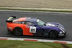 Photographie d'une voiture de grand tourisme violette, orange, noire et blanche, vue de haut et de profil, sur une piste.