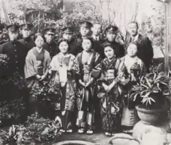 Photo noir et blanc de plusieurs femmes en tenue japonaise traditionnelle, avec derrière elles des hommes en uniforme sombre ou en costume.