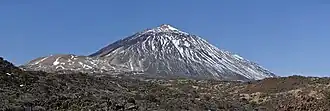 Photo du volcan conique partiellement enneigé avec un dôme marqué à sa gauche et un terrain rocheux au premier plan.