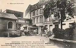 Carte postale en noir et blanc. Voitures attelées sur une place bordée de maisons et d'immeubles.