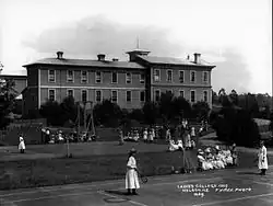 Photographie en noir et blanc représentant des femmes en train de joueur au tennis devant un bâtiment en arrière-plan.