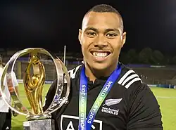 Un homme de face avec un vêtement noir portant un trophée sportif dorée.