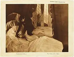 Une lobby card avec une photographie au centre, et de petites légendes en dessous. L'image représente un homme vêtu de noir qui se penche vers le lit blanc d'une jeune femme endormie.