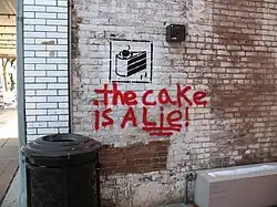 Photo du mur d'une ruelle, sur lequel sont graffés la phrase "The cake is a lie!" en rouge, et l'icône de gâteau de Portal