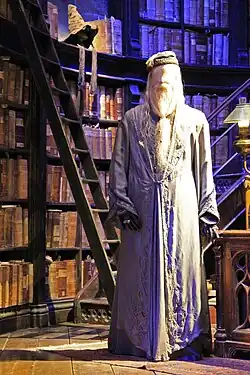 Un pantin debout vêtu d'une longue robe masculine grise, placé devant une grande bibliothèque de livres anciens sur un mur circulaire.