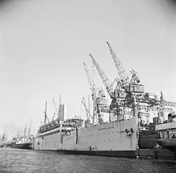 Photographie en noir et blanc d'un navire amarré à un quai, à côté de grues métalliques.