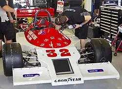 Photo d'une monoplace rouge et blanche dans un garage. Deux mécaniciens travaillent sur cette voiture.
