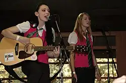 Deux jeunes filles en train de chanter (une brune à gauche jouant de la guitare et une blonde à droite). Elles portent des gilets rose foncé sans manches sur des chemisiers blancs, avec des cravates vertes visibles