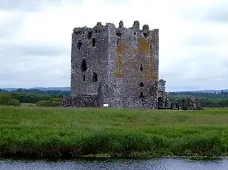 Un château au centre, entouré d'herbe et d'eau, le ciel et des établissement à l'arrière plan