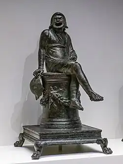 Acteur assis, jambes croisées, sur un socle et portant un masque à la bouche très largement ouverte.