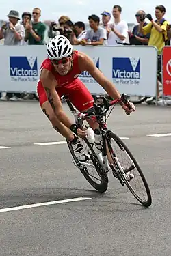 Un homme portant un casque et un maillot de cyclisme rouge est penché sur un vélo de route, en train de prendre un virage serré sur une route goudronnée. Il participe au triathlon des Jeux du Commonwealth 2006 à St Kilda, en Australie.