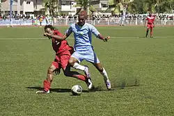 Photographie d'une scène d'un match de football : un joueur en bleu bouscule un autre joueur rouge pour le ballon.