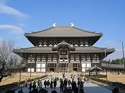 Photo couleur d'un imposant édifice religieux bouddhique en bois, à deux étages. Arrangement de poutres et de panneaux blancs, avec deux toitures de tuiles grises, sur fond de ciel bleu. Au premier plan, quelques dizaines de personnes se tiennent debout, dans l'allée qui mène aux marches de l'entrée du bâtiment.