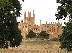 Photo d'un bâtiment en pierre ocre de grande taille, coiffé de nombreuses tourelles et caché derrière un rideau d'arbres