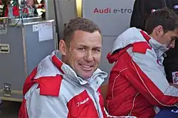 Un homme brun, vu de face, regarde à sa droite, et sourit. Il est vêtu d'un manteau gris et rouge.