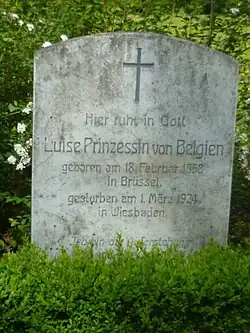 Une petite stèle en pierre claire au sommet arrondi est placé derrière un massif de verdure.