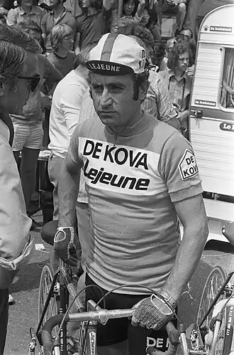 Photographie en noir et blanc montrant un cycliste tenant son vélo à côté de lui, en conversation avec un autre homme.