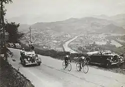 Photographie en noir et blanc de deux cyclistes suivis par une voiture dans l'ascension d'une route de montagne.