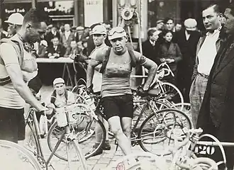 Photographie en noir et blanc d'un cycliste se tenant debout, les fesses appuyées sur le cadre de son vélo, devant la foule.