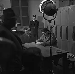 Scène d'intérieur lors d'un tournage, éclairée par des projecteurs : Fernandel et Terzieff sont assis de deux côtés différents d'une table, avec la mine grave. Au fond de la pièce un agent de police en uniforme semble en faction.