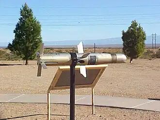Photographie en couleurs d'un missile gris métallisé reposant sur un support noir devant une pancarte, deux arbres en arrière-plan