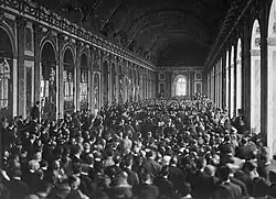 Photographie de la très longue et large Galerie des glaces où une foule innombrable se tient debout autour de personnes assises en groupe sur des chaises.